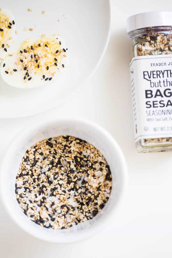 A small white bowl of everything bagel seasoning next to a jar of the Trader Joe's version. And a hard boiled egg with some of the seasoning sprinkled on the cut side.