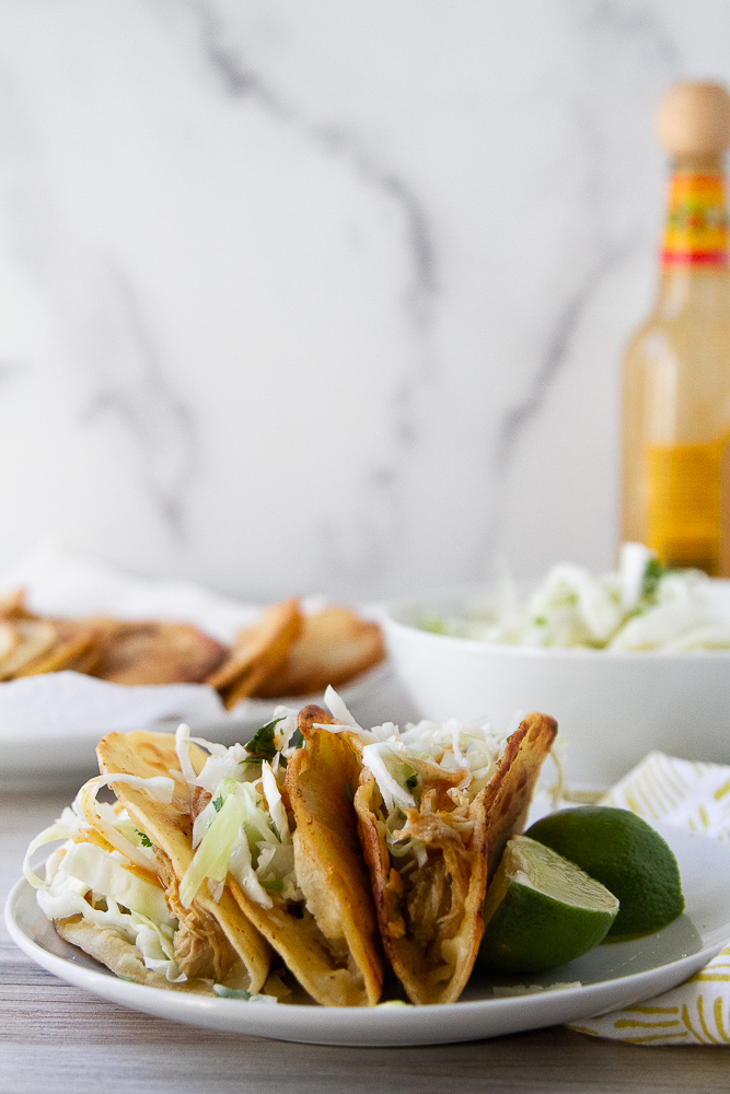 Side view of crispy chicken tacos on a plate with cabbage slaw and lime.