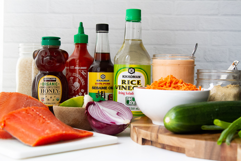 Ingredients laid out for the salmon rice bowls: fresh salmon fillets, cucumber, carrots, onion, lime, garlic, green onions, spicy mayo, rice, and ingredients for the sauce (honey, soy sauce, sesame oil, sriracha, rice vinegar, ginger, salt, and sugar.)