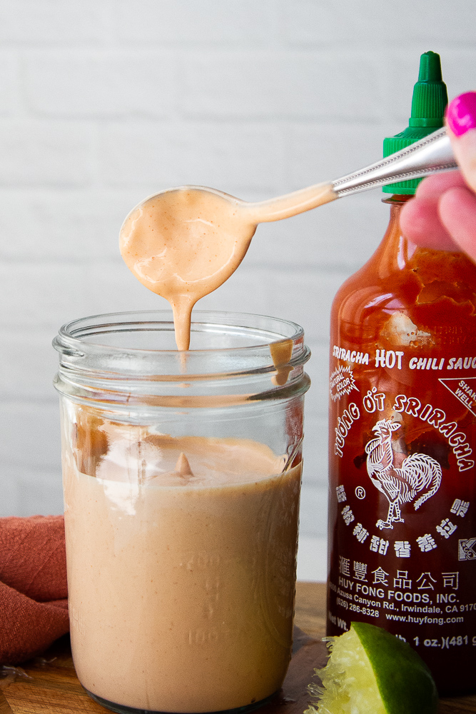A mason jar with spicy mayo covered spoon hovering over the top with mayo dripping off. Sitting next to a bottle of sriracha sauce.