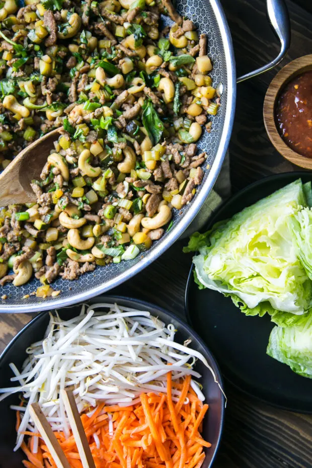 Finished Thai Chicken Lettuce Wrap filling on a table with iceberg lettuce wedges, a bowl of sambal oelek, and shredded carrots & mung bean sprouts ready to assemble.