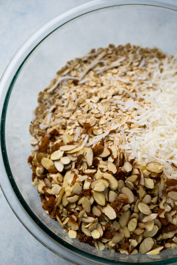 A large, clear mixing bowl with oats, coconut, and sliced almonds.