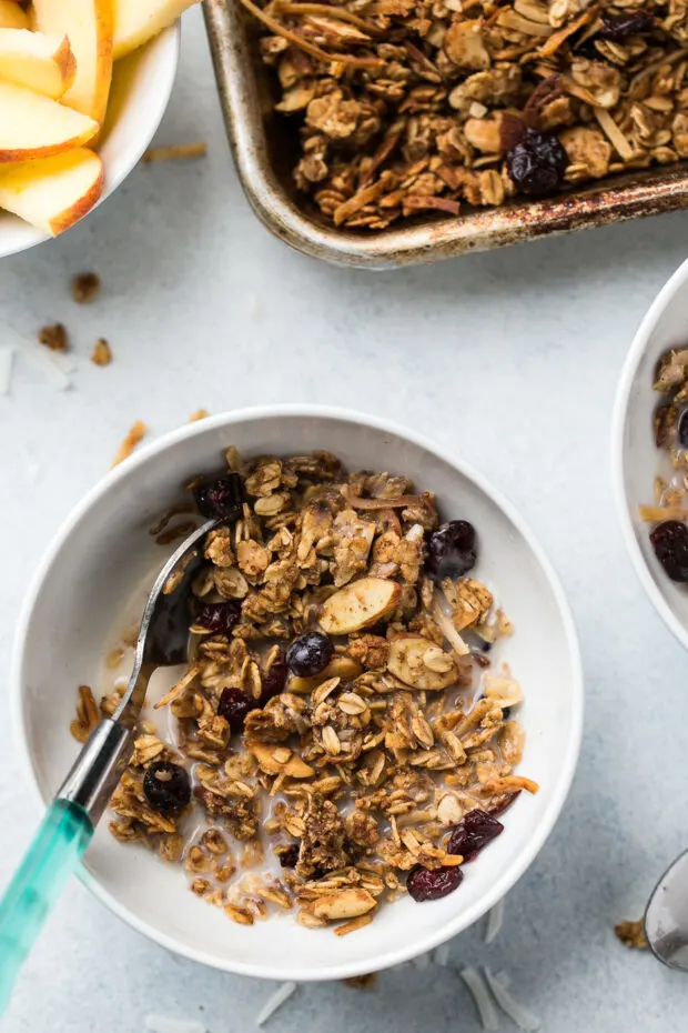 Top down flat lay of a rimmed baking sheet with baked granola and two bowls of granola with milk.