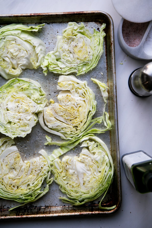 Six cabbage steaks on a rimmed baking sheet. They have been sprinkled with salt, pepper, and drizzled with oil and are ready to broil.
