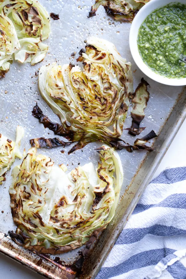 Freshly broiled cabbage steaks on a sheet pan with a small bowl of pesto ready for serving.