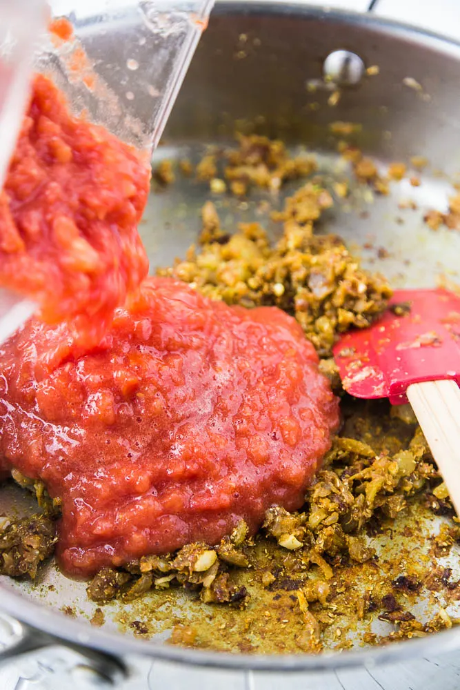 Blended tomatoes being poured from a blender pitcher into the skillet. Spices have been added to the onions, too.