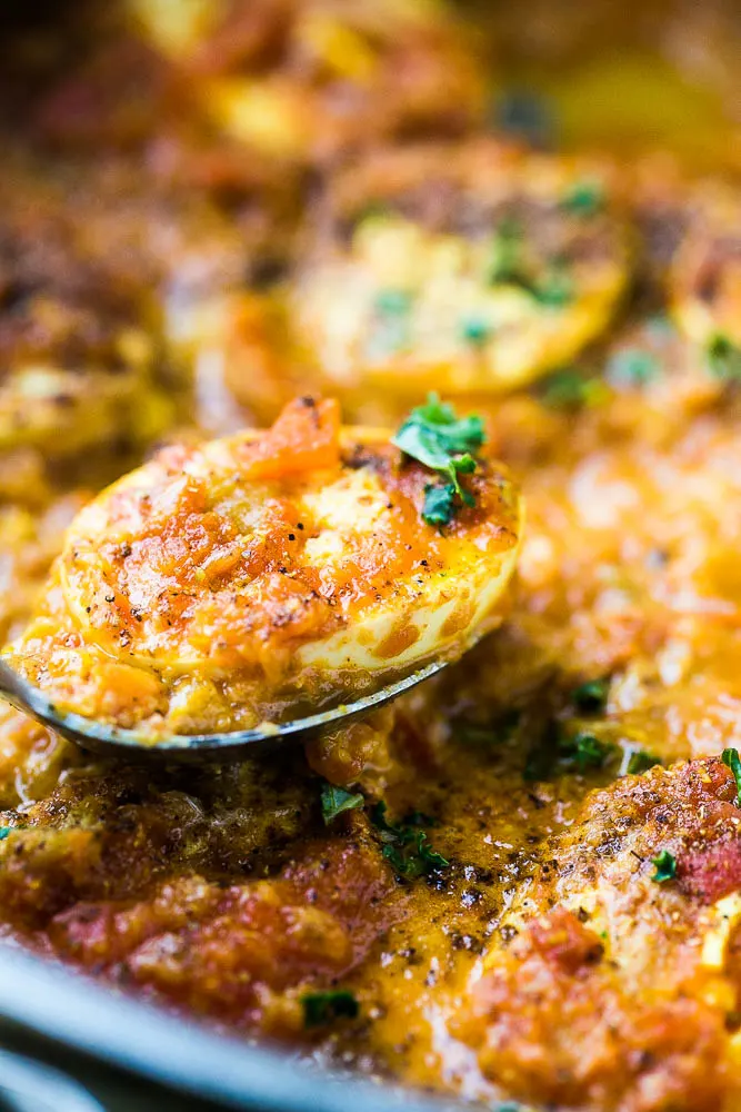 An egg being scooped out of a skillet containing egg curry.