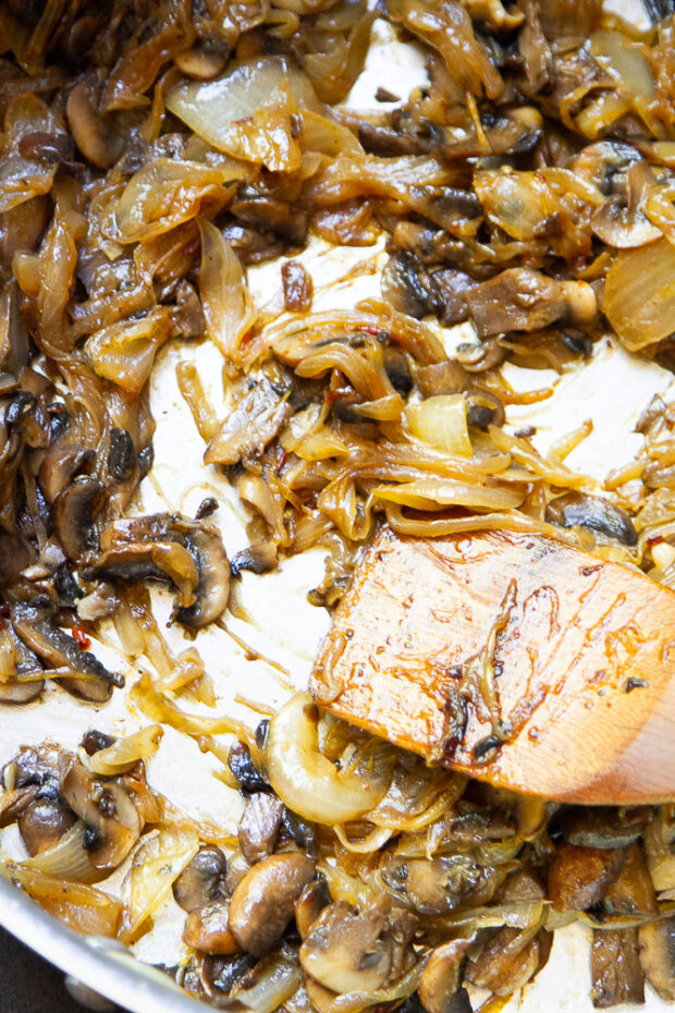 Shot of completely caramelized onions mixed with mushrooms being stirred with a wooden spatula.