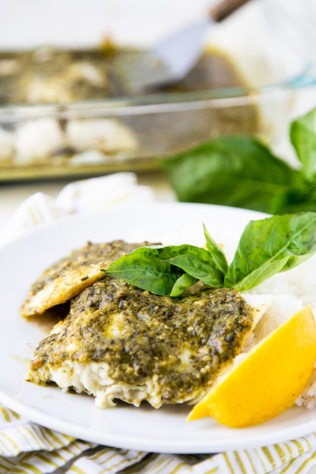 A pretty white plate with pesto-covered baked cod with white rice, a lemon wedge, and a sprig of fresh basil.