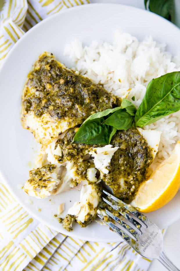 Baked Cod covered in pesto on a white plate with white rice and a lemon wedge.