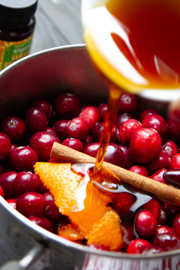 Adding maple syrup to a saucepan along with cranberries, cinnamon stick, and orange peel.