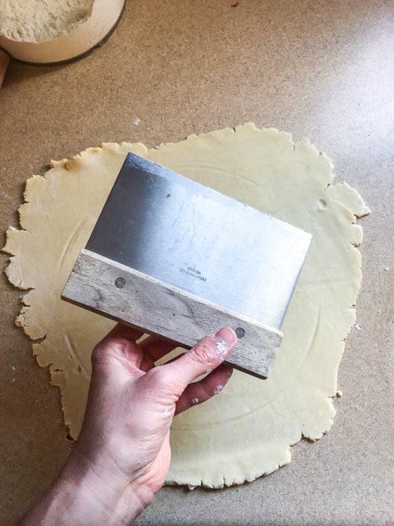 View of a pastry scraper held over the rolled out pie crust dough.