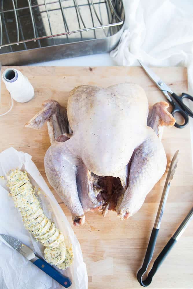 Brined turkey ready to be prepped for the oven. On the cutting board with the turkey is the herb butter cut into "coins", tongs, kitchen twine, shears, and a sharp knife.