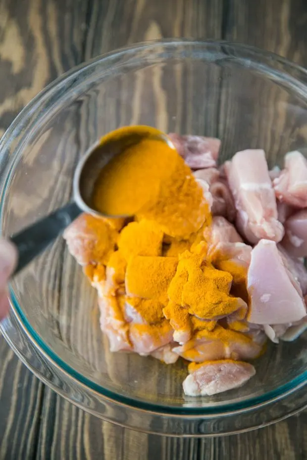 Chopped chicken pieces in a clear class bowl with turmeric being sprinkled over the top.
