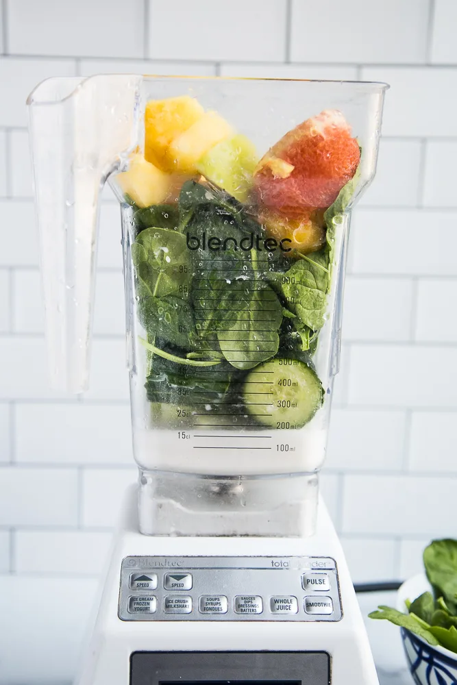 View from the side of the blender showing the green smoothie ingredients -- lots of leafy greens, cucumber, celery, milk, pineapple, apple, and orange.