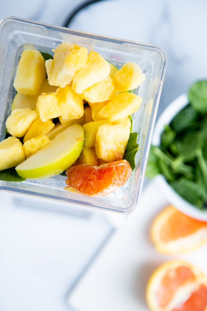 Top down view of blender pitcher. Frozen pineapple and apple and orange slices are added on top of the leafy greens.