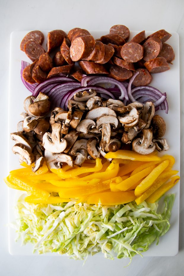 Easy Sausage Skillet Meal using prepped vegetables and sausage slices. Cabbage, bell peppers, mushrooms, onion, andouille sausage on a cutting board.