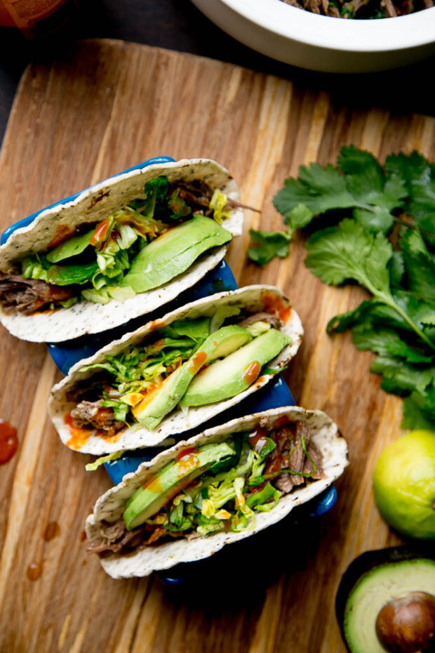 Three green chile shredded beef tacos topped with lettuce, avocado, & hot sauce.