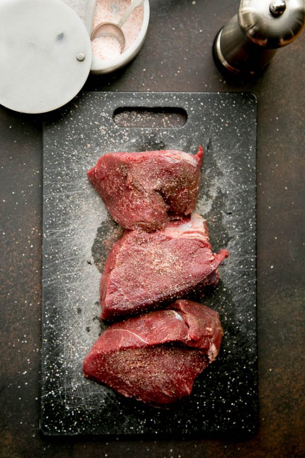 A beef roast cut into three big pieces sitting on a black cutting board.