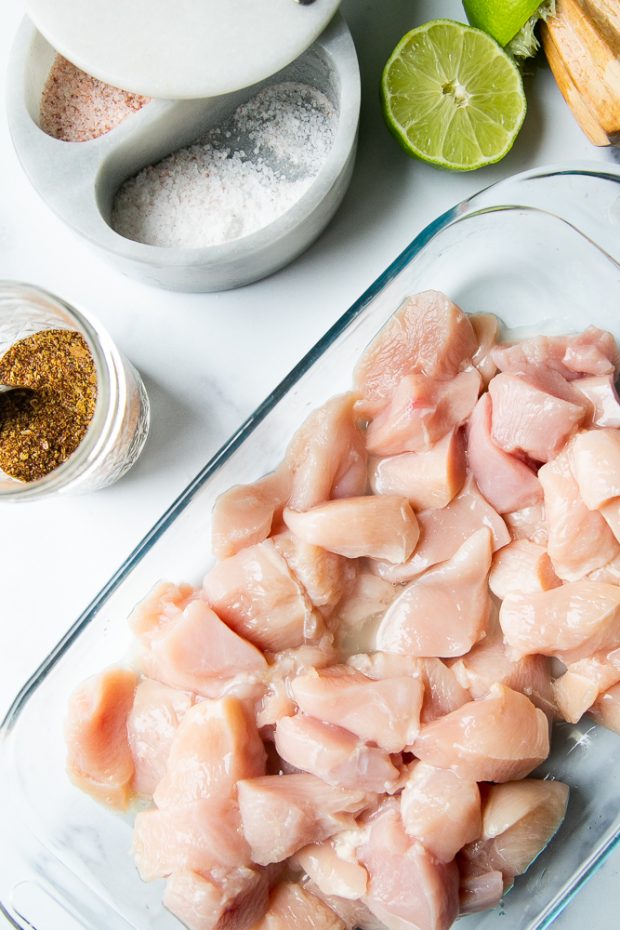 A glass dish with small pieces of raw chicken. Alongside sit some limes, salt, and taco seasoning