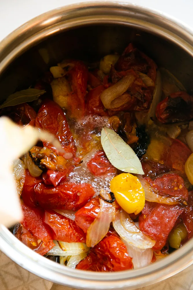 Roasted tomatoes, onions, & garlic in a pot. Chicken broth is being poured in to prepare for simmering.