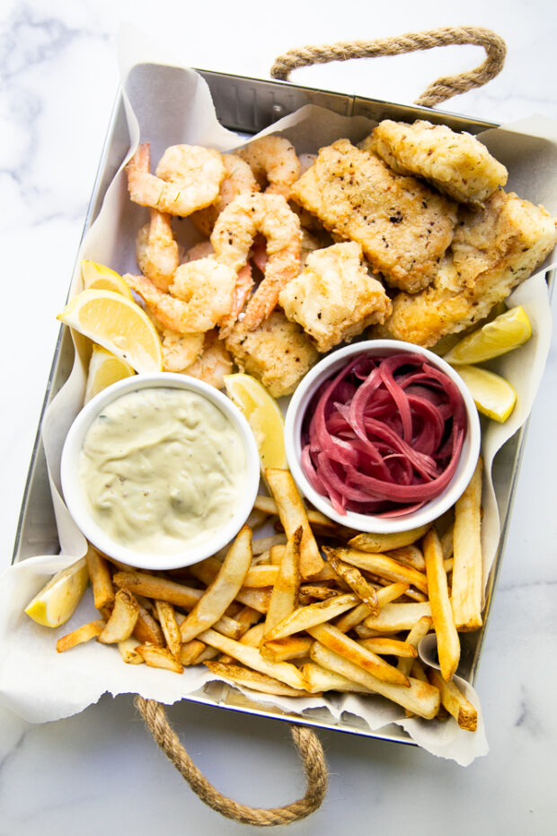 This Aruba-inspired fish fry is one of our favorite meals and memories from a fun family trip. Image shows a tray with fried pieces cod & shrimp, french fries, garlic aioli, and pickled red onions.