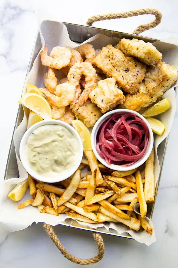 This Aruba-inspired fish fry is one of our favorite meals and memories from a fun family trip. Image shows a tray with fried pieces cod & shrimp, french fries, garlic aioli, and pickled red onions.