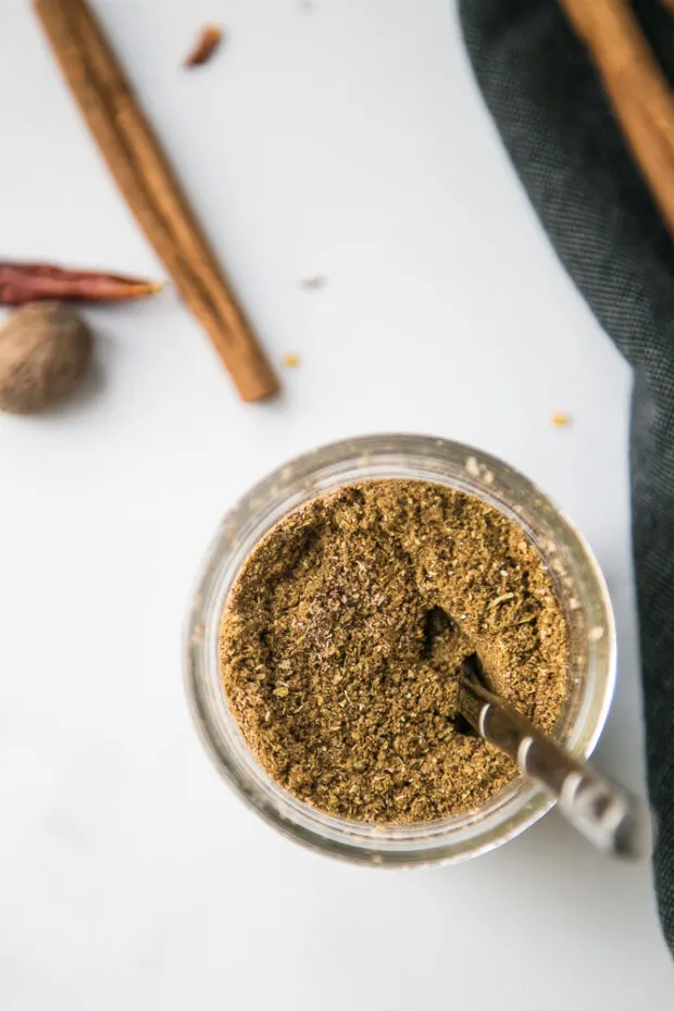 Ground up spices blended to make garam masala. Spice blend is in a glass mason jar surrounded by bits of cinnamon sticks and chili peppers.