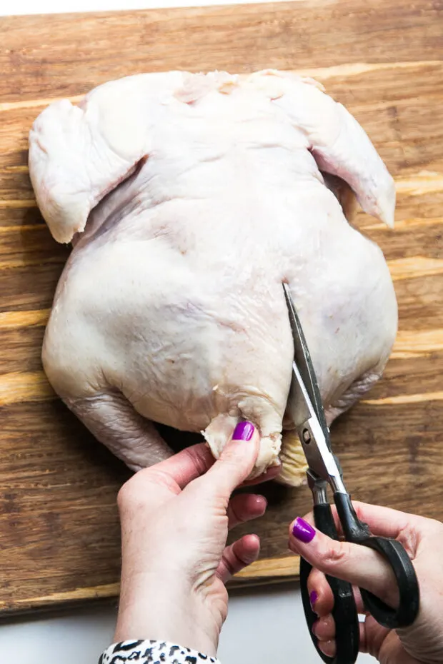 Whole raw chicken laying breast-side down on a cutting board. Find the base of the backbone and cut slightly to the right.