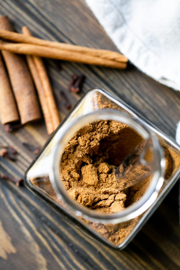 Homemade pumpkin pie spice in a clear glass jar. Cinnamon sticks and whole cloves are on the table next to the jar.