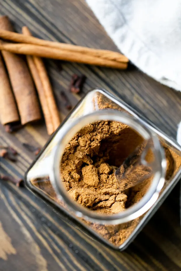 Homemade pumpkin pie spice in a clear glass jar. Cinnamon sticks and whole cloves are on the table next to the jar.