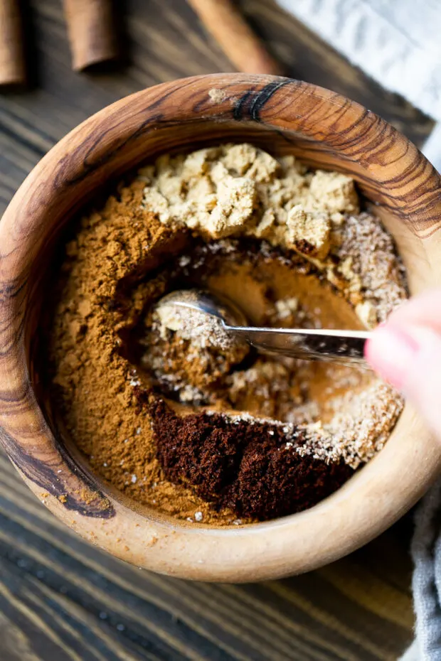 The ingredients for pumpkin pie spice are being stirred by a measuring spoon in a small wooden bowl.