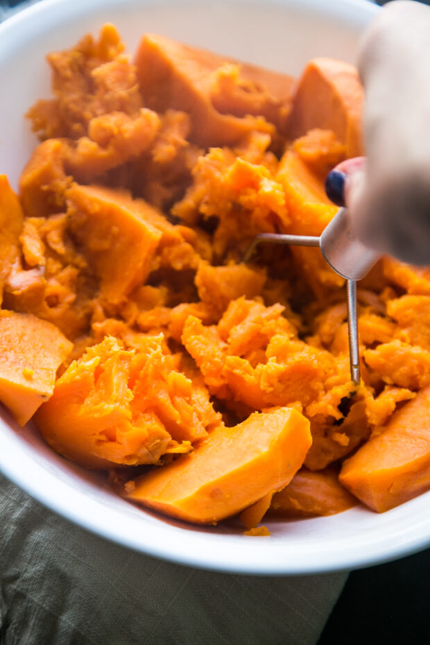 Cooked sweet potatoes are being mashed in a white serving bowl with a small potato masher.