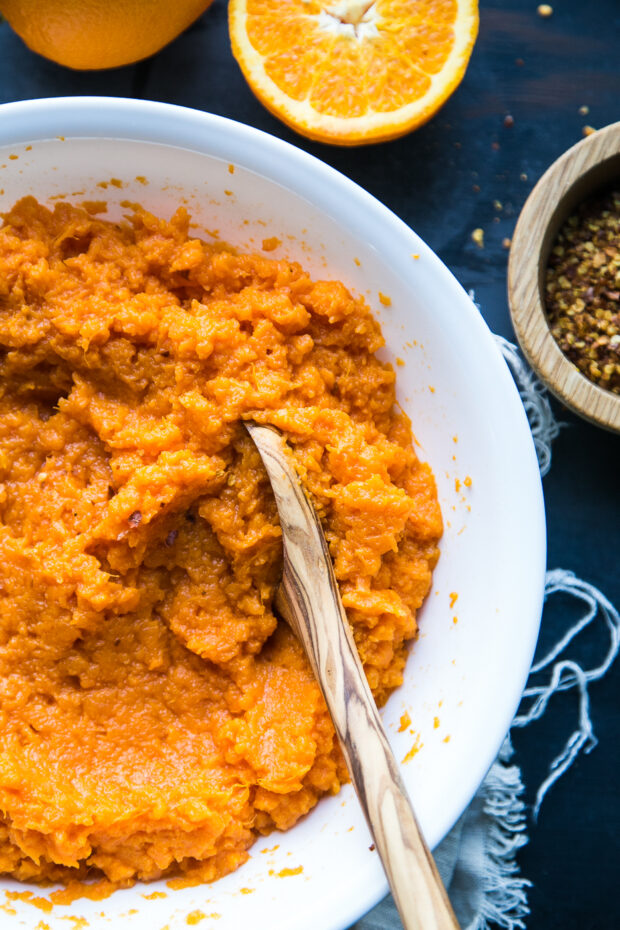 A large white bowl of mashed sweet potatoes with a wooden serving spoon. The bowl is next to a smaller wooden bowl with crushed red pepper flakes and an orange that has been cut in half.