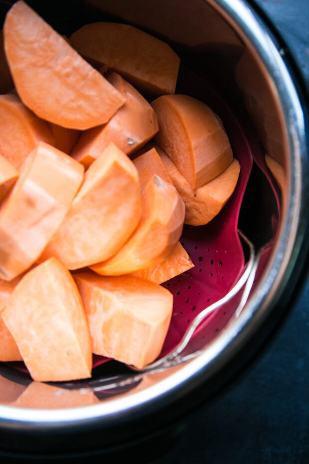 View looking into an Instant Pot. The pot is filled with large, uncooked pieces of sweet potato. The sweet potatoes are sitting on a silicone steam basket on top of the wire rack.