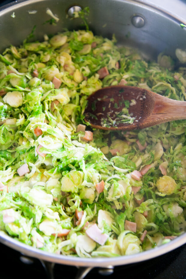 Shredded Brussels & ham in a skillet cooking and stirred with a large wooden spoon.