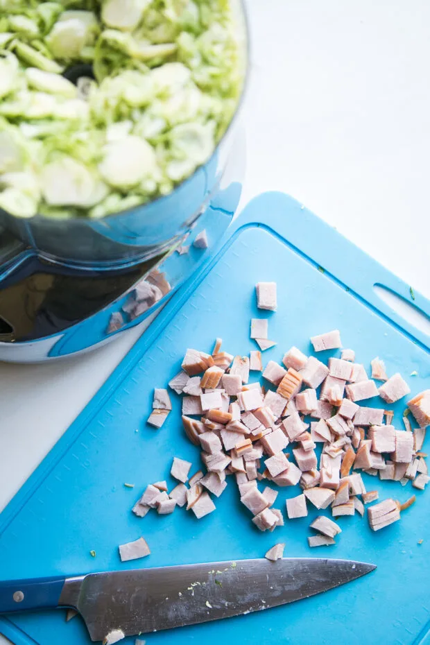 Shredded Brussels sprouts in a food processor next to a blue cutting board with diced deli ham.