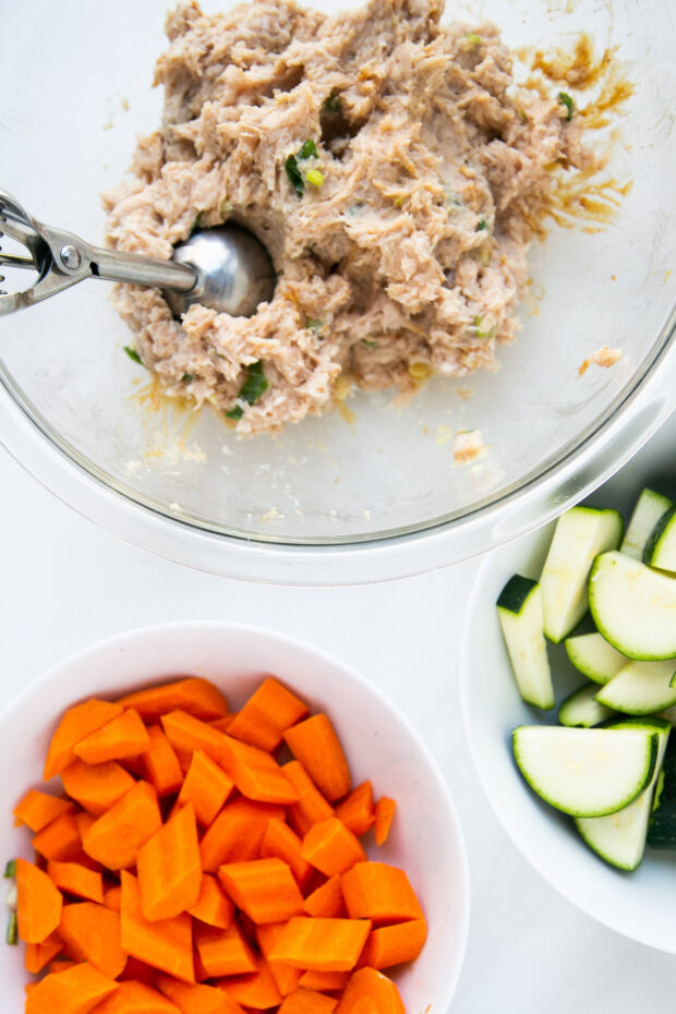 Large bowl with the raw ginger chicken meatball mixture and a cookie scoop sticking out of it. Two smaller white bowls containing chopped zucchini and carrots.