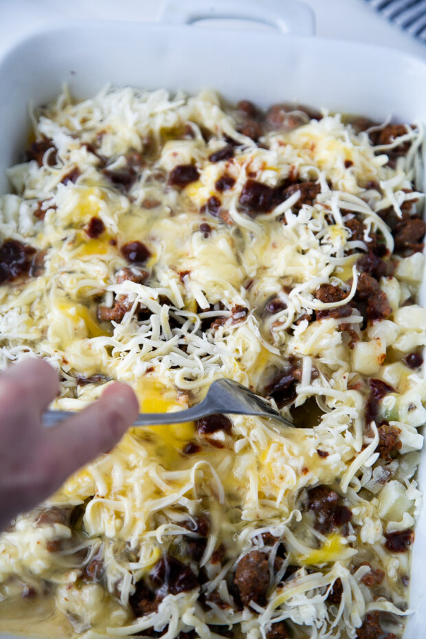 Uncooked breakfast casserole in a white baking dish. A fork is stirring the mixture around a little.