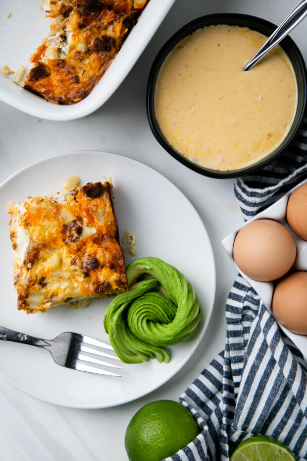 A square of the breakfast casserole sits on a white plate next to an avocado rose. Nearby are some raw brown eggs, a bowl of hollandaise sauce, and the casserole dish.