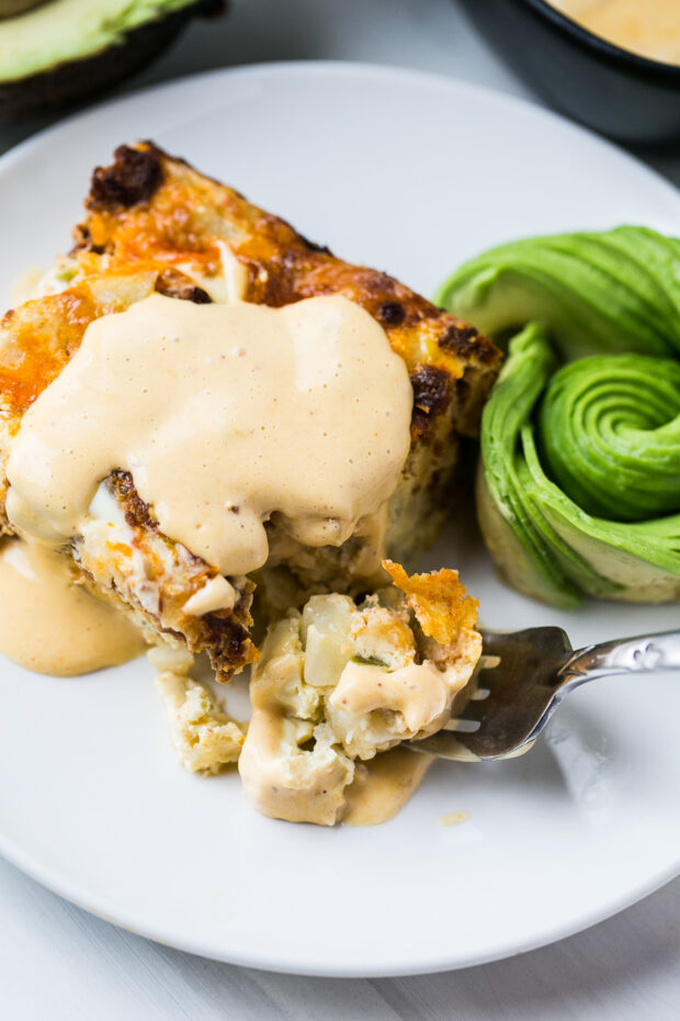 A square of chorizo hashbrown casserole on a white plate topped with hollandaise sauce. An avocado rose sits next to the breakfast casserole.
