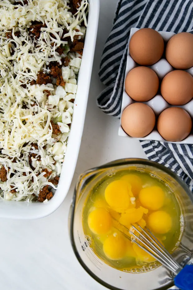 Overhead view: White casserole dish with frozen hashbrowns, cooked chorizo, and pepperjack cheese. Next to it is a black and white striped towel with a container of eggs on top. A liquid measuring cup containing cracked eggs sits on the bottom.