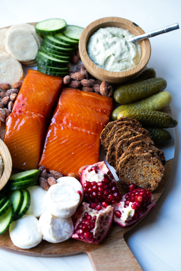 Hot smoked salmon on a cutting board with dill pickles, spiced almonds, pomegranates, dips, and crackers.