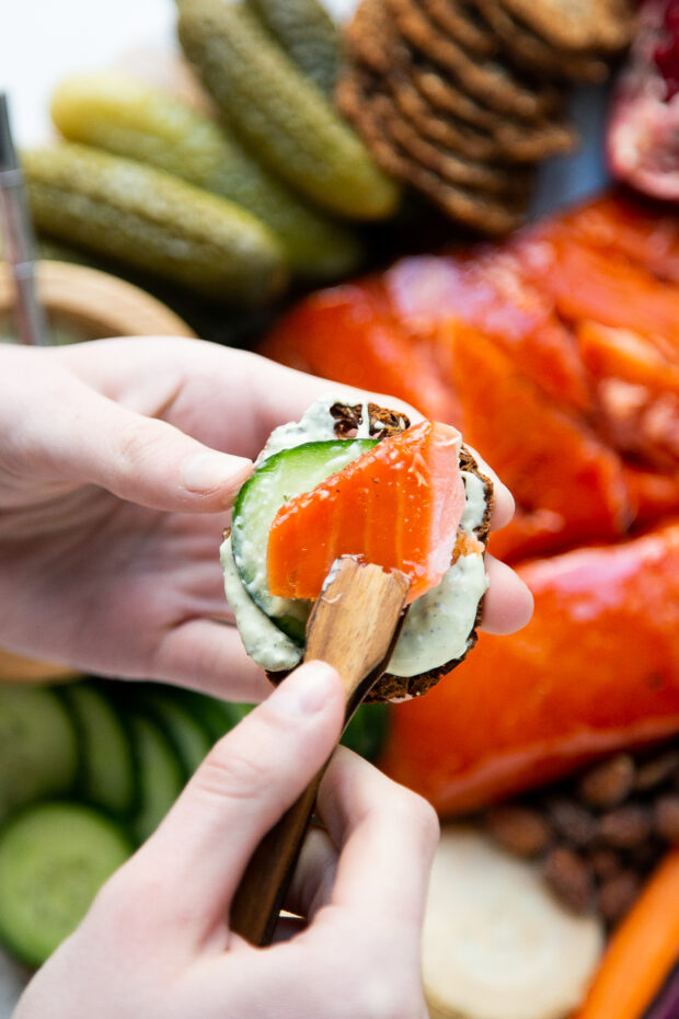 A mini salmon crostini on a whole grain cracker with green goddess dip and cucumber.