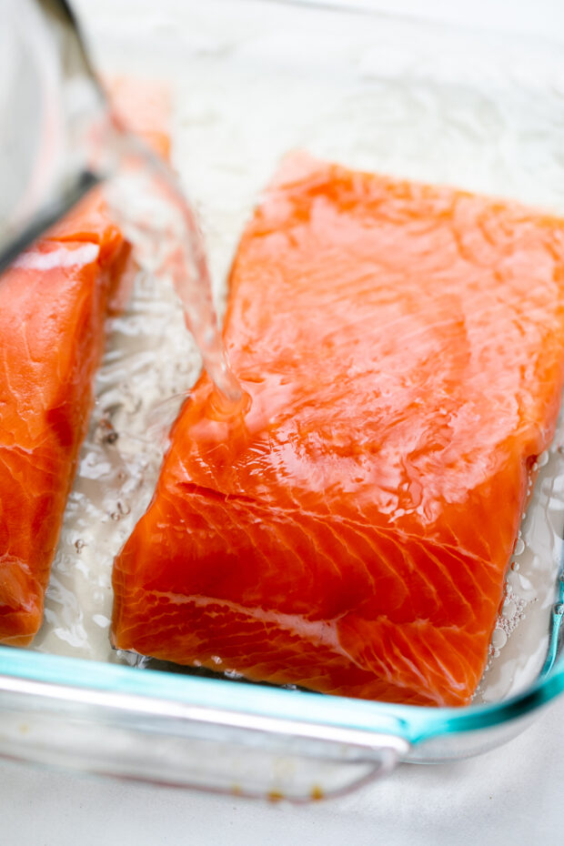 Raw fish fillets in a glass baking dish. Clear brine is being poured over the salmon.