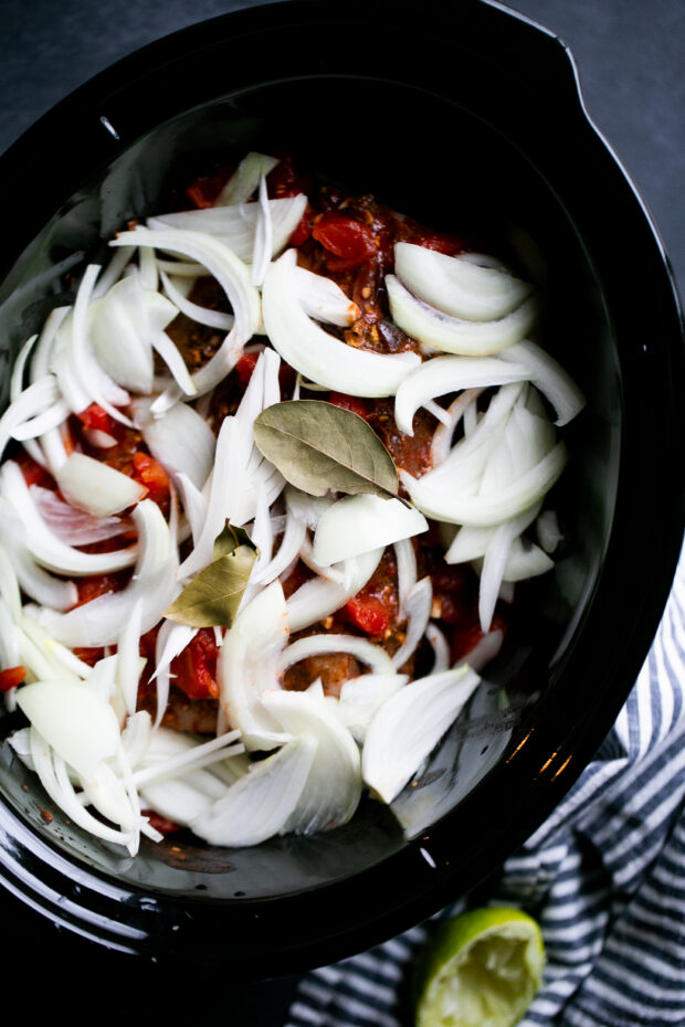 Spice covered roast in a slow cooker topped with raw onions, diced tomatoes, and bay leaves.