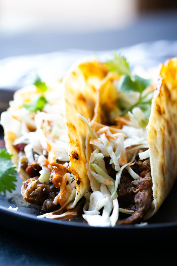 Barbacoa Beef Tacos with shredded cabbage in a gluten-free tortilla. Garnished with hot sauce and fresh cilantro sprigs.