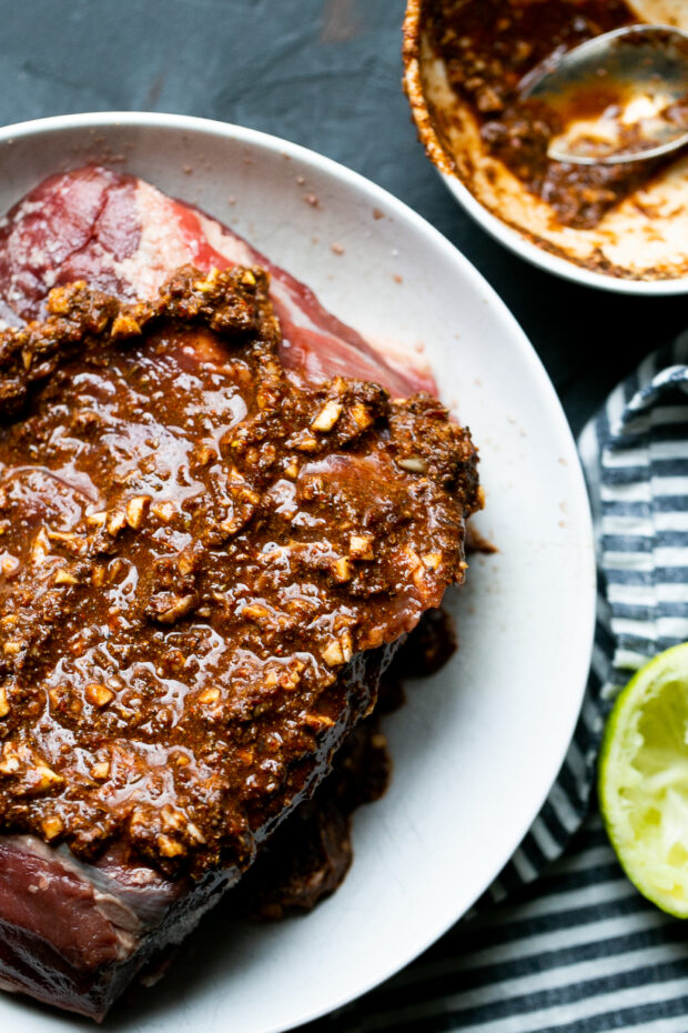 Raw beef top round roast in a white bowl. Roast is covered in the chipotle lime spice paste.