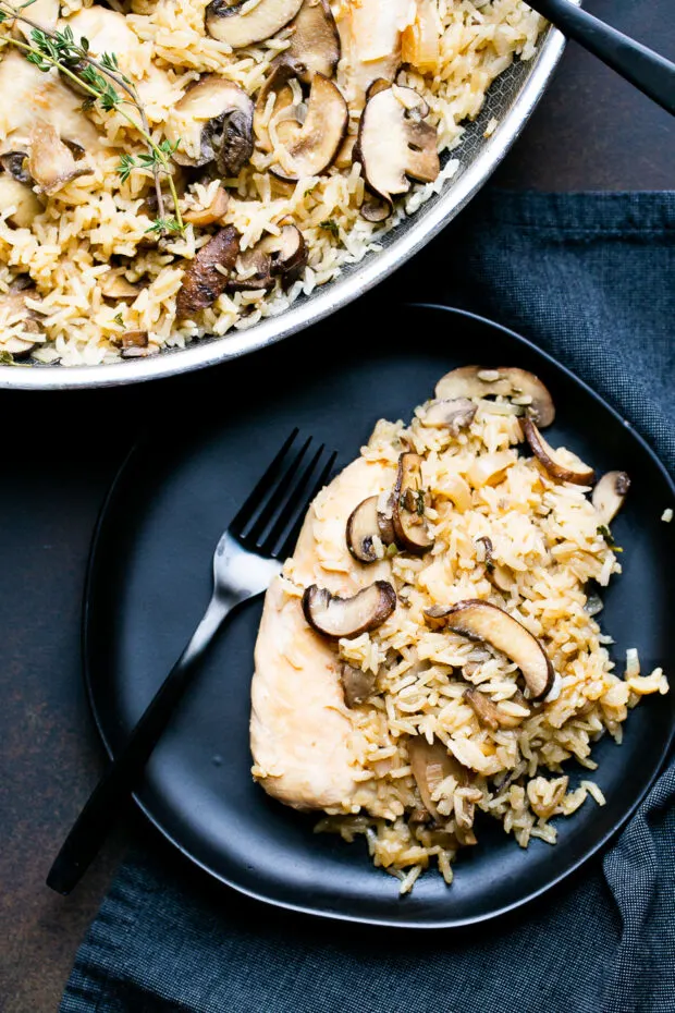 A black plate with a chicken breast and some mushroom rice next to a black fork.
