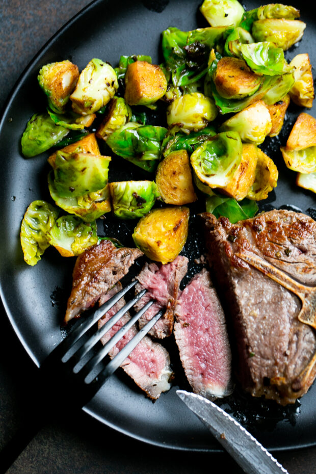 Partially sliced lamb loin chop on a black plate with sauteed Brussels sprouts. 